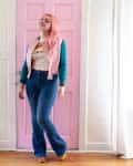 Woman with pink hair in casual outfit standing by a pink door, next to a hanging chair with a cushion.