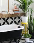 Clawfoot bathtub with black and white geometric tiles, wooden shelf, potted plants, and a striped towel.