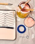 Paint supplies on a drop cloth, including a roller, tray with pink paint, brush, tape, and open paint can.