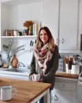 Woman in a cozy kitchen holding a mug, with wooden countertops, white cabinets, and shelves with jars and books.