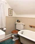 Small bathroom with a pedestal sink, clawfoot tub, and toilet with a wooden seat under a sloped ceiling.