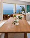Open-plan living and dining area with ocean view, wooden table, white benches, potted plants, and modern decor.
