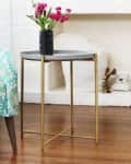 Gray side table with gold legs, black vase with pink flowers, next to a patterned turquoise chair on wooden floor.
