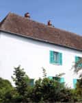 White house with teal shutters and a steep roof, surrounded by lush greenery under a clear blue sky.