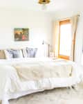 Bright bedroom with a large bed, white bedding, two bedside lamps, a wall painting, and a window with beige curtains.