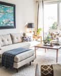 Bright living room with beige sectional sofa, blue throw, wooden coffee table, and large window with sheer curtains.