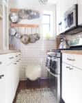 Galley kitchen with white cabinets, stainless steel appliances, hanging pots, and a small window.
