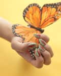 Hand with green and red nail art holding a painted orange butterfly on a yellow background.