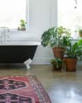 Freestanding black bathtub with claw feet, surrounded by potted plants and a pink patterned rug on concrete floor.