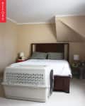 Bedroom with beige walls, wooden bed, gray pillows, white bedding, and a large pet crate at the foot of the bed.