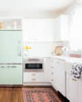 Bright kitchen with mint green fridge, white cabinets, wooden floor, and decorative rug.