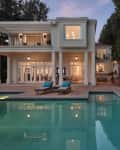 Two-story house with large windows, pool, patio, lounge chairs, and potted plants at dusk.