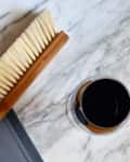 Wooden-handled brush on a marble surface next to a glass of dark liquid and a gray mat.