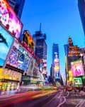 Times Square at dusk with vibrant billboards, bustling crowds, and blurred lights from passing traffic.