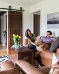 Couple sitting on a brown leather sofa with a golden retriever, next to a wooden chest and yellow tulips in a vase.