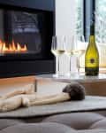 Cozy living room with a fireplace, wine bottle, and glasses on a table, knit gloves, and a window view of snowy trees.