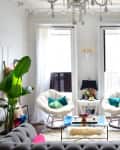 Bright living room with two white chairs, teal pillows, a gray sofa, large plant, wall-mounted TV, and chandelier.