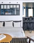 Living room with a gray sofa, black and white pillows, wooden coffee table, and a view into a kitchen with bar stools.