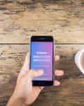 Hand holding a smartphone with Instagram login screen next to a cup of coffee on a wooden table.