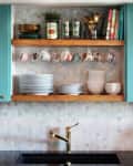 Open kitchen shelf with hanging floral teacups, stacked plates, bowls, cookbooks, and a gold faucet below.