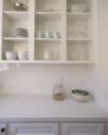 Open kitchen shelves with white dishes, mugs, glassware, and a countertop with a jar and nested bowls.