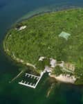Aerial view of a lush green island with a dock, sandy beach, and tennis court surrounded by water.