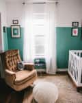 Nursery with brown armchair, white pouf, crib, and teal accent wall. Framed art and sheer curtains on window.