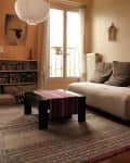 Cozy living room with a beige sofa, black coffee table, striped runner, bookshelf, and large window with sheer curtains.