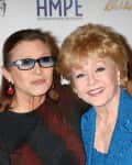 Two women smiling, one in sunglasses and earrings, the other in a blue outfit, posing together at an event.