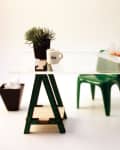 Glass desk with green sawhorse legs, a small plant, mug labeled "Ann," sandwich, clock, and green chair.