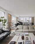 Spacious living room with a grand piano, beige sofa, black leather chairs, and large windows overlooking the city.