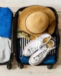 Open suitcase with blue towel, gray hoodie, straw hat, sunglasses, striped shirt, and white sneakers on wooden floor.