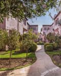 Mediterranean-style courtyard with pink stucco buildings, lush greenery, and a winding stone path.