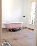 Pink clawfoot bathtub on wooden floor in a partially renovated bathroom with white walls and a window.