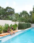 Two women sitting on orange cushions by a pool, surrounded by lush greenery and palm trees.