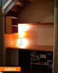 Under-stair nook with wooden desk, shelves, and a lit table lamp.