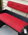 Red cushioned metal bench with a tag, placed against a beige siding wall.