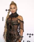 Woman in a sheer black beaded gown with a long braided ponytail, standing against a backdrop with geometric patterns.