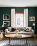Living room with gray sofa, geometric pillows, wooden coffee table, and green walls, featuring framed art and white curtains.