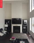 Living room with a TV above a fireplace, flanked by black shelves, a gray rug, and a black coffee table with magazines.