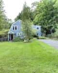Blue house with red door, surrounded by lush greenery and white hydrangeas, set in a spacious yard.