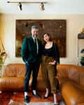 Two people standing in a stylish living room with a brown leather sofa, abstract wall art, and a patterned rug.