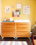 Wooden dresser with changing pad, colorful wall art, and striped rug in a yellow nursery.