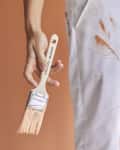 Hand holding a paintbrush with orange paint, wearing white pants splattered with paint.