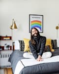 Person sitting on a bed with yellow and black pillows, rainbow artwork, and brass wall lamps in a cozy bedroom.