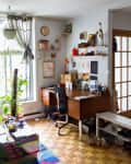 Home office with wooden desk, chair, shelves with decor, plants by window, and colorful rug on parquet floor.