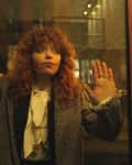 Woman with curly hair and a white blouse stands behind a glass door, hands pressed against it.