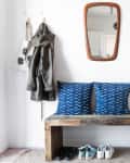 Entryway with wooden bench, blue patterned cushions, hanging plants, mirror, coat, and shoes neatly arranged below.