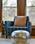 Blue armchair with orange pillow, silver pouf, cactus in terracotta pot, and wooden side table by large windows.
