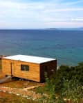 Wooden cabin overlooking a calm sea with a yacht, surrounded by greenery and distant mountains.